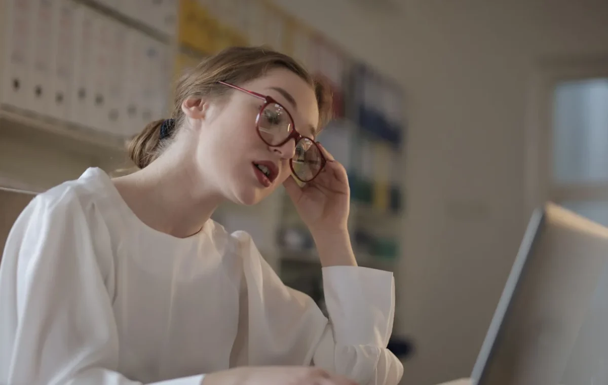 A focused woman with eyeglasses works on her laptop in a modern office setting.