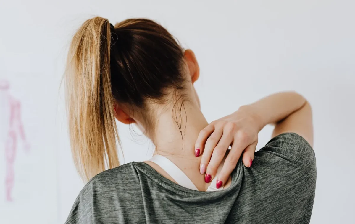 Back view of slim blond female in casual wear with manicure indicating pain spot on back while sitting in front of white wall with paper drawing in clinic