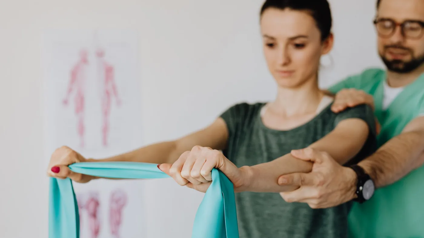 Crop osteopath in uniform and eyeglasses supporting shoulder and reached arm of young woman with elastic tape in doctor office in hospital on blurred background