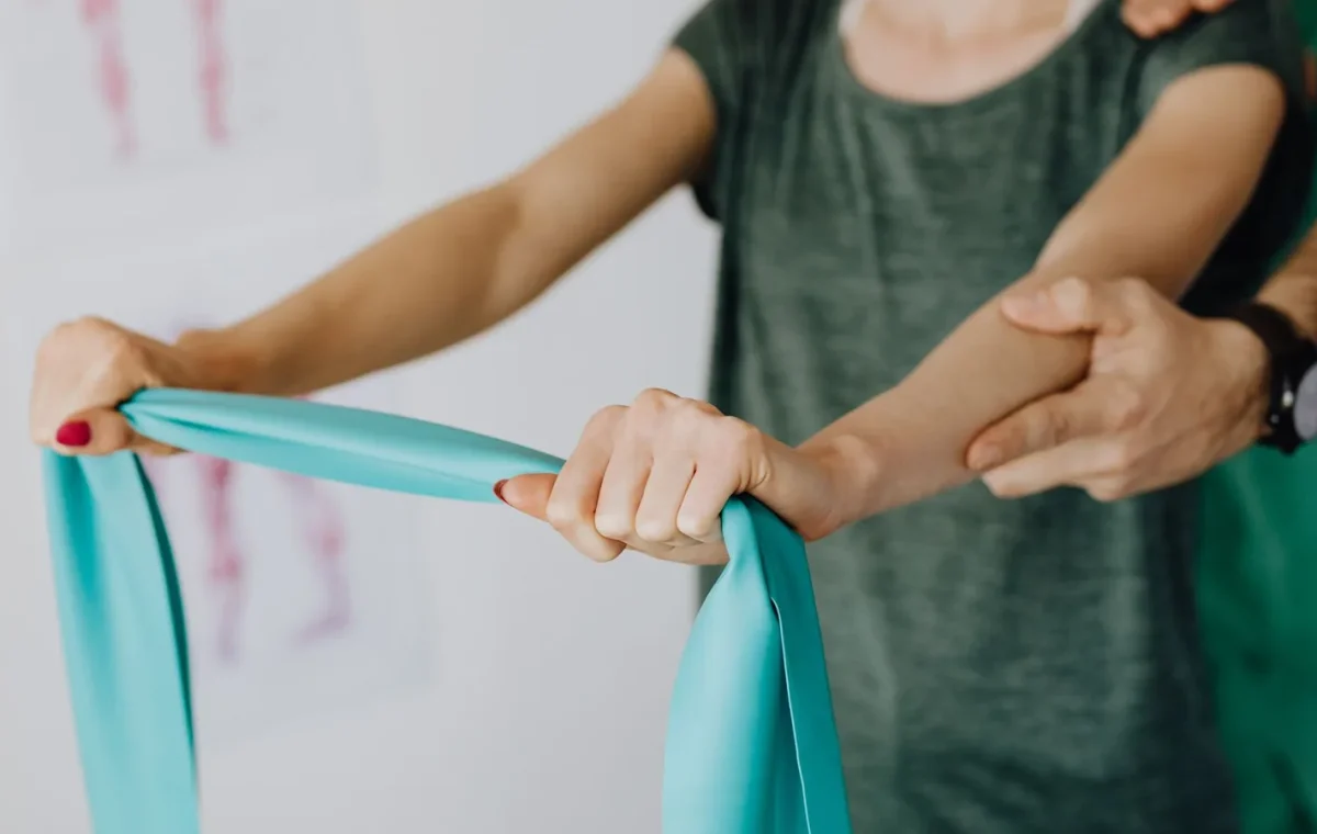 Crop unrecognizable orthopedist in uniform and wristwatch supporting shoulder and reached arm of faceless female patient with resistance band in doctor office