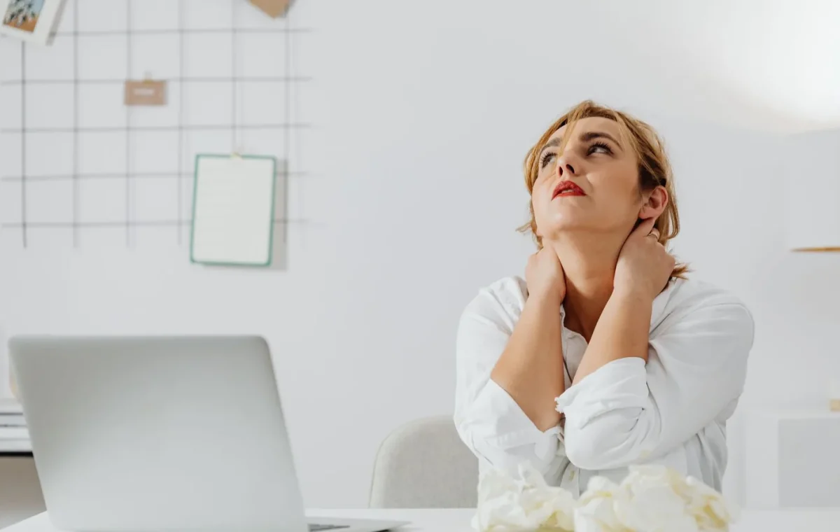 A stressed woman in white long sleeves sits at a desk with a laptop, looking upwards in an office.