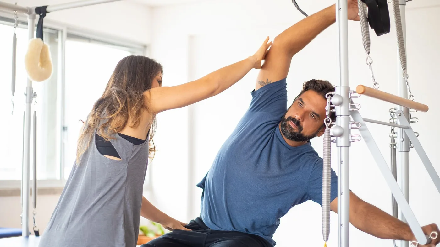 Trainer guides client on reformer machine during pilates session in a bright gym setting.