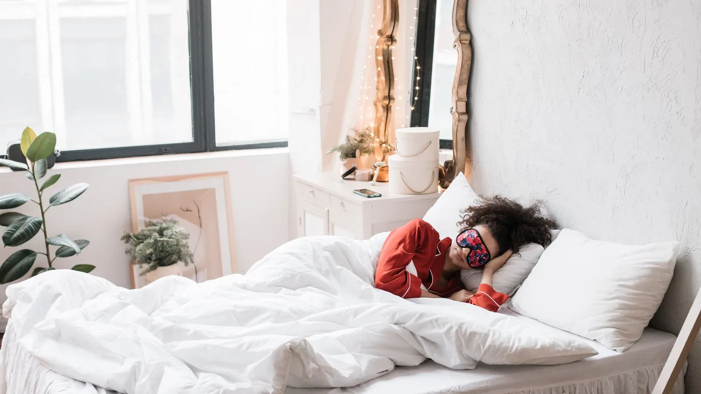 Woman wearing a sleep mask, resting on a cozy white bed surrounded by indoor plants in a bright bedroom.