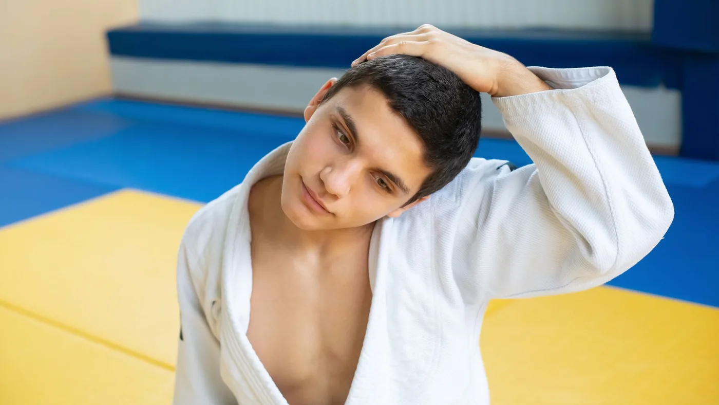 A young man in a judo uniform stretches his neck in a brightly colored gym setting, expressing focus and concentration.