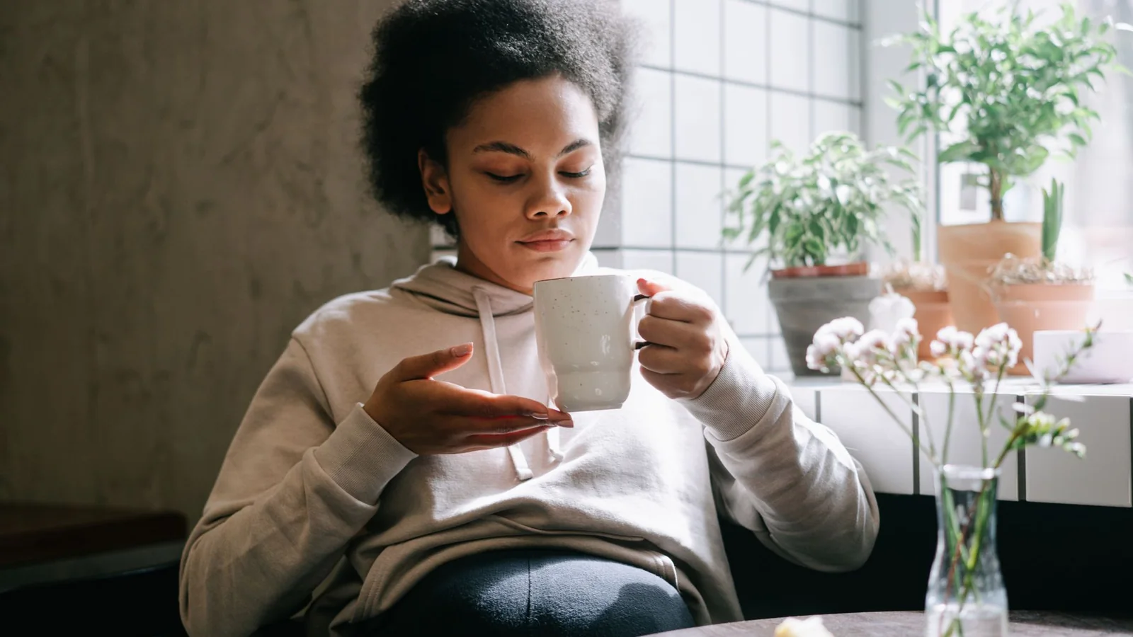 Woman enjoying a peaceful moment sipping tea by a window with indoor plants.