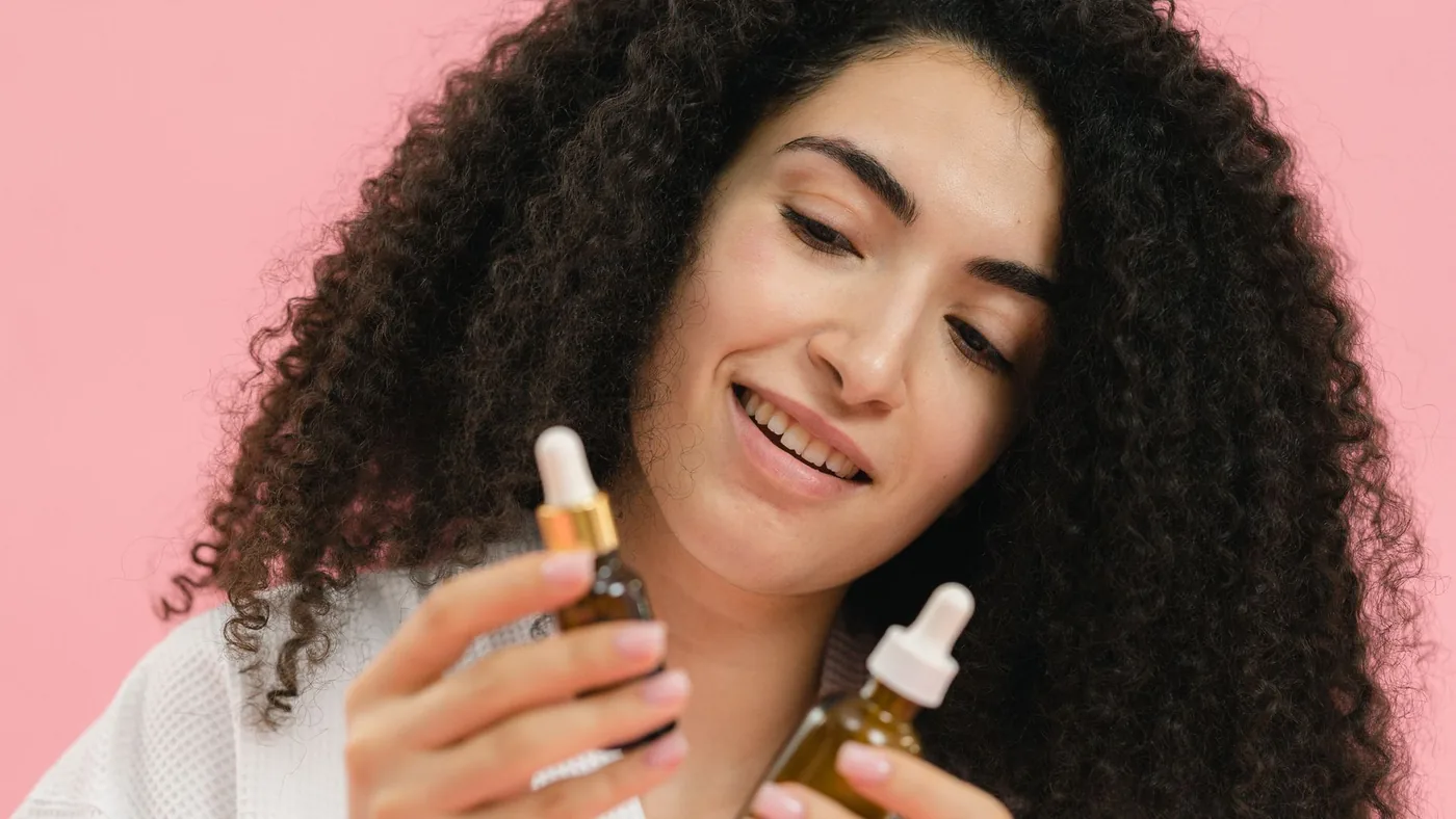 A young woman analyzes skincare bottles, set against a pink backdrop.