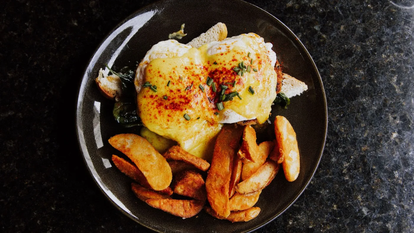 Delicious Eggs Benedict with creamy sauce and crispy potato wedges served on a dark plate.