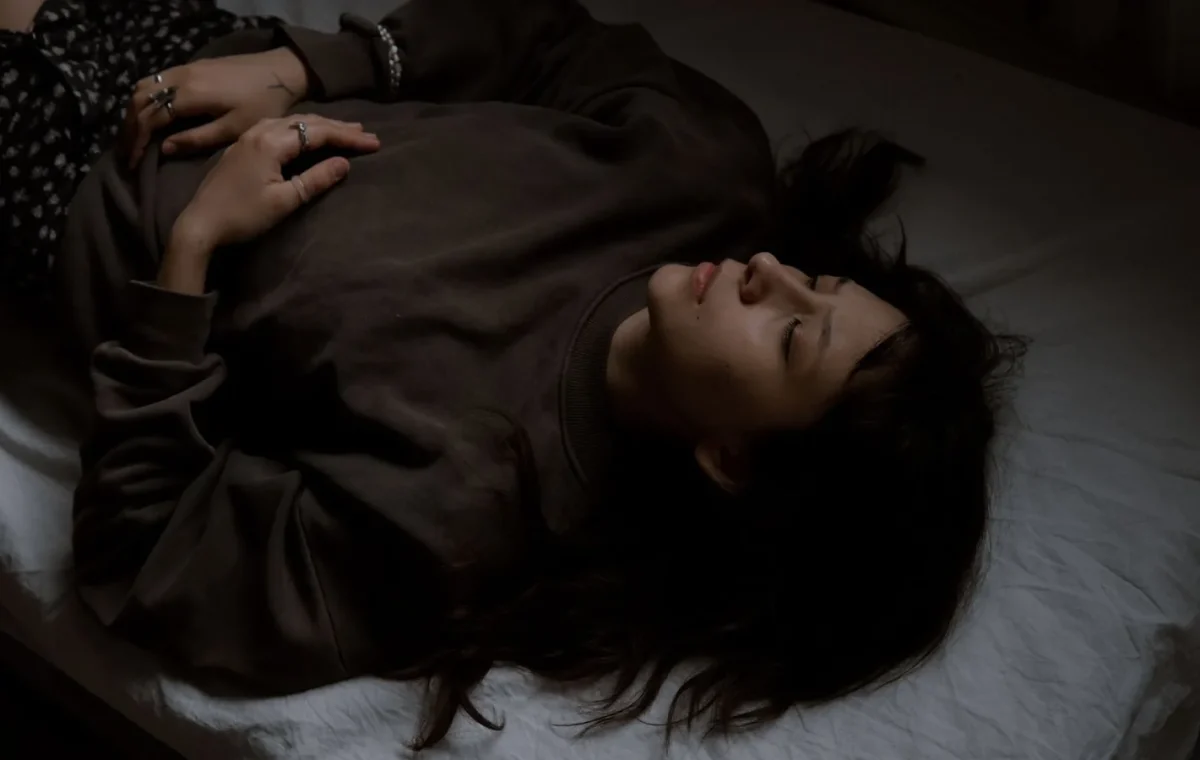 Serene young woman with closed eyes lying on a bed in a dimly lit room.