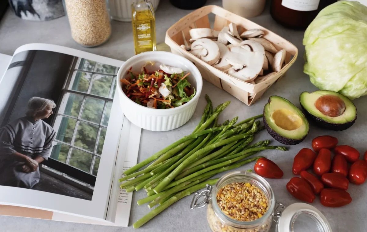 A selection of fresh vegetables and a cookbook in a kitchen setting, promoting healthy cooking.