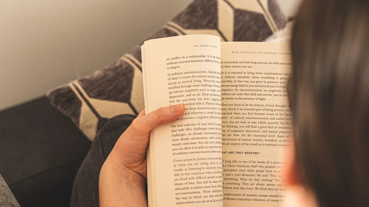 A person enjoys a relaxing reading session indoors, holding an open book on a comfy couch.
