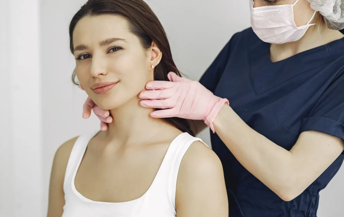 A medical practitioner in scrub suit performs a neck examination on a smiling woman in a clinic.