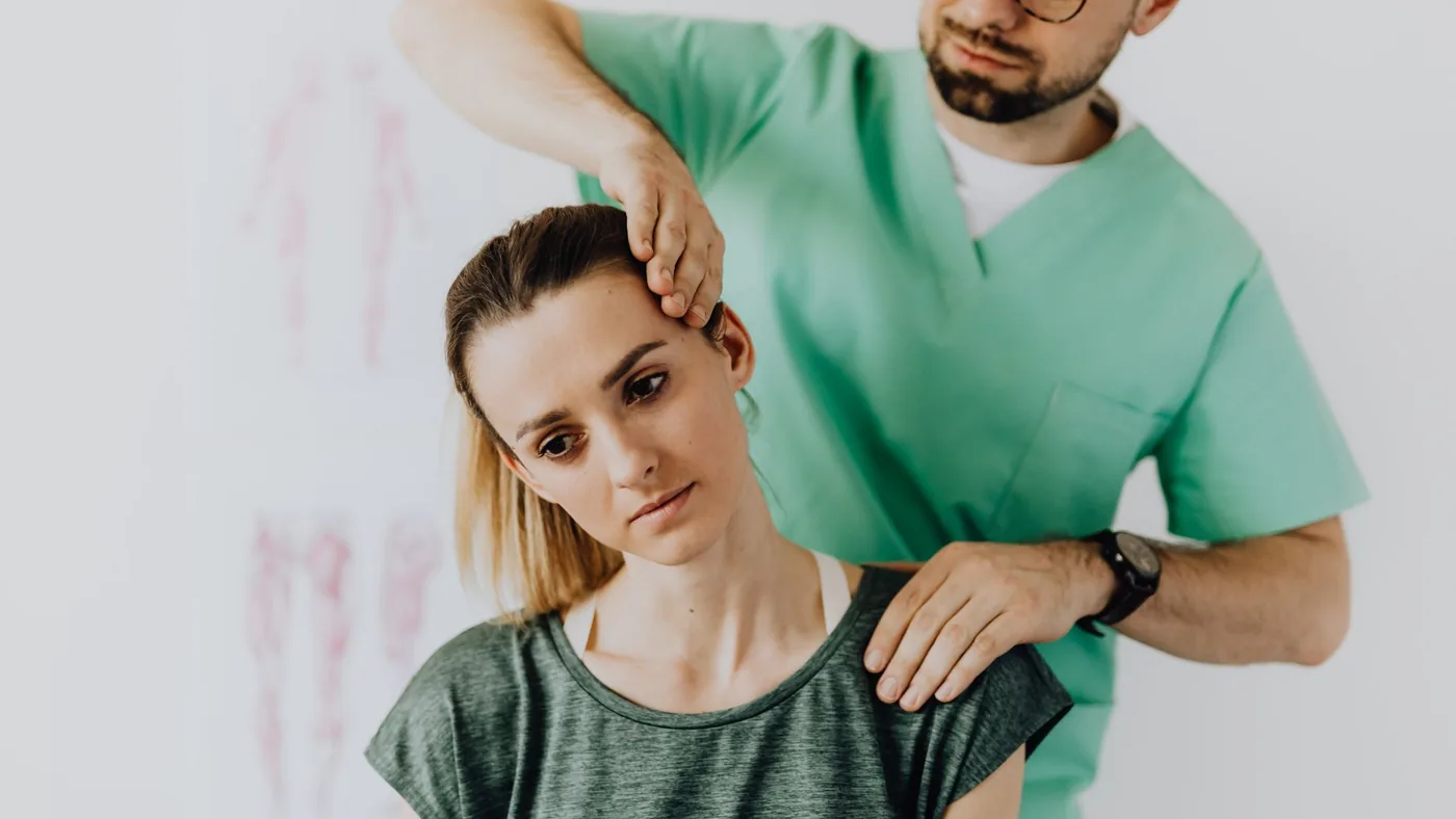 Crop osteopath in uniform and eyeglasses standing with closed eyes while examining neck of young thoughtful female in casual wear in hospital