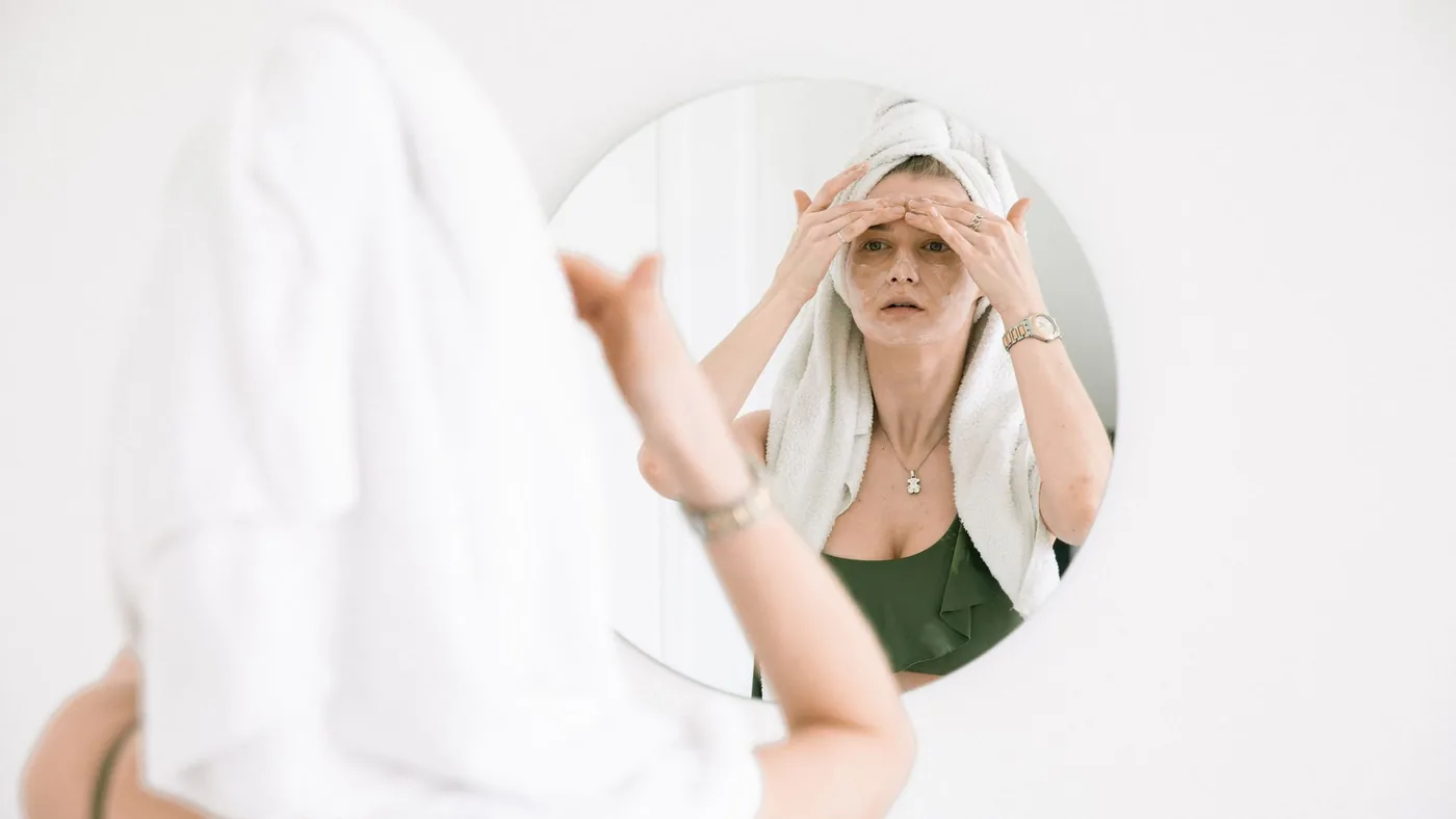 Woman with towel on head applies skincare, reflects self-care routine in mirror.