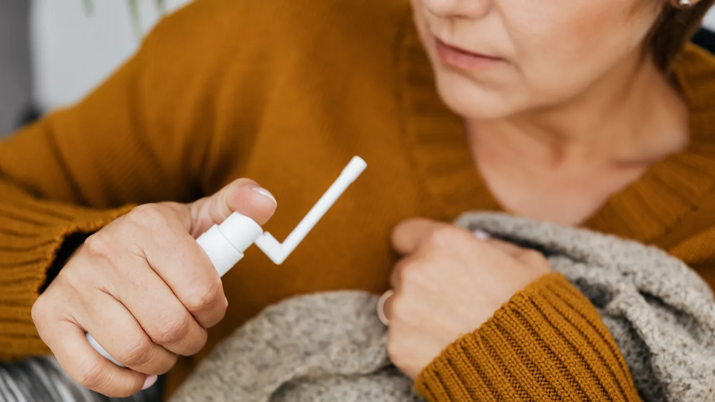 Adult woman using a mouth spray for relief indoors, wrapped in a cozy sweater.