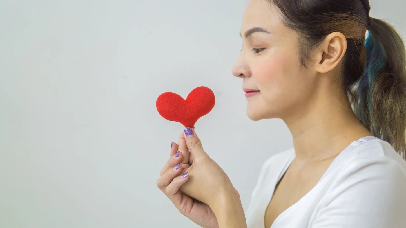 Side view of calm female in ponytail with red heart in hand and standing on white background in light room