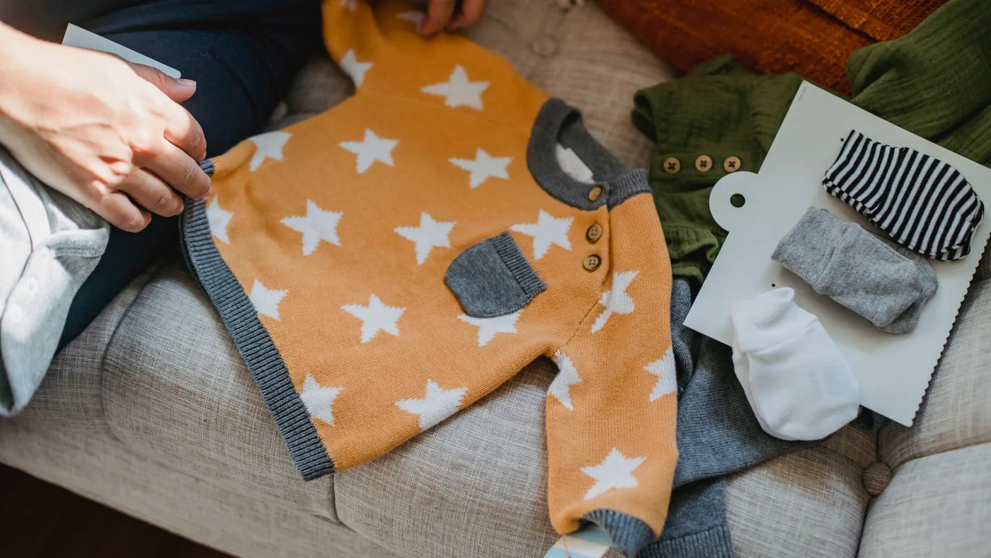 High angle of crop faceless parent sitting on sofa and showing new garment for baby