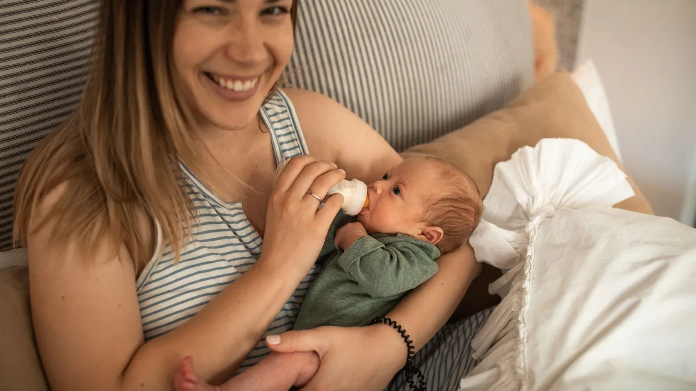 A mother lovingly feeds her newborn with a bottle while smiling at the camera.