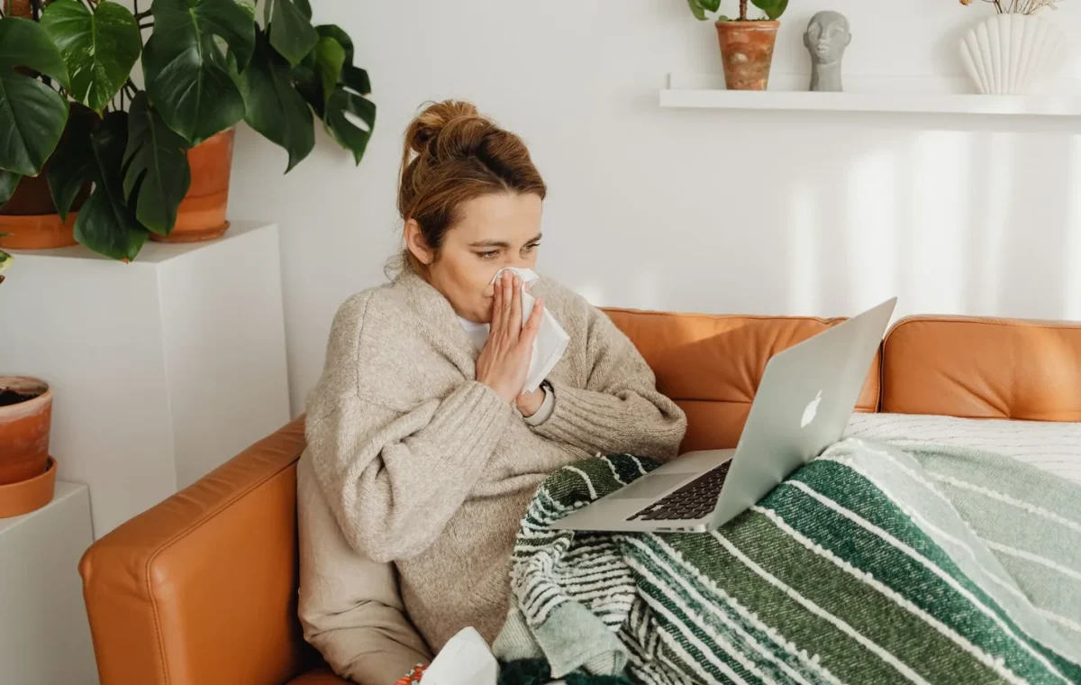 Woman in cozy sweater using laptop on sofa, surrounded by indoor plants, while blowing nose.