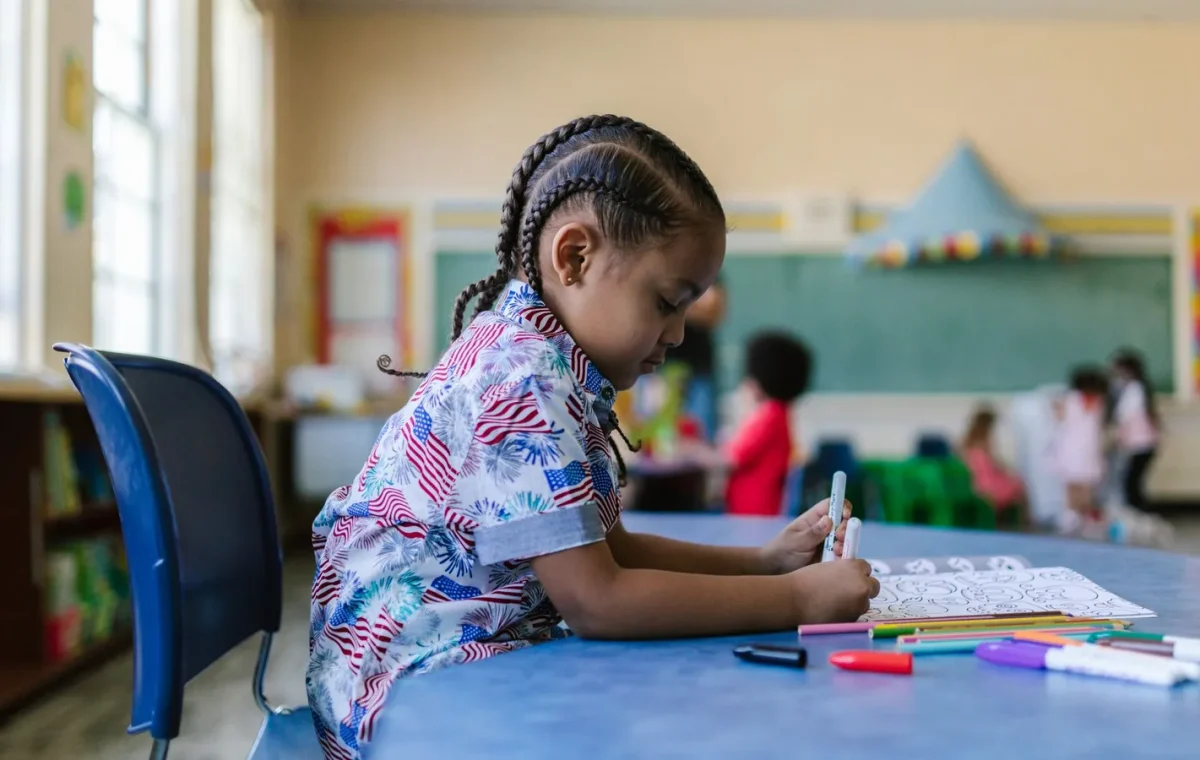 A young child with braids engaged in coloring at a classroom desk surrounded by school supplies.