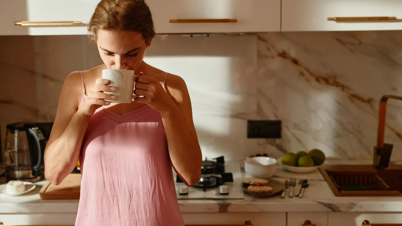 A woman enjoys a hot beverage in a sunlit kitchen, creating a tranquil morning scene.